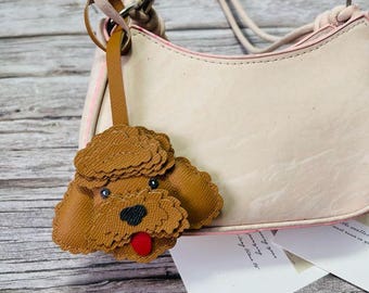 Puede incluir: Un bolso rosa claro con una correa a juego y un charm con forma de caniche marrón. El bolso tiene una forma curvada con ribete rosa. El charm tiene una lengua roja y ojos negros.