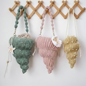 May include: Three crochet seashell-shaped bags in seafoam green, blush pink, and gold hang on a wooden rack. The bags have beaded straps and are embellished with a starfish, a white flower, and pearls.