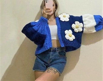 May include: A blue and white knitted cardigan with large white crocheted flowers. The cardigan has a cropped length and long sleeves.