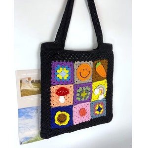 May include: Black crochet tote bag with colourful square patches featuring a smiley face, mushroom, rainbow, and other designs. The bag has two long handles and is made of a textured, woven material. A magazine is visible in the background.