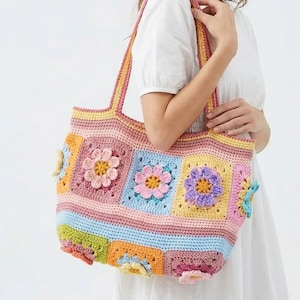 May include: A colorful, handmade crochet tote bag with a floral design. The bag features a patchwork pattern with squares of pink, blue, yellow, and green, each adorned with a crocheted flower. The bag has matching pink handles.