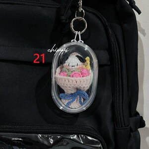 May include: A clear, oval keychain featuring a crocheted Snoopy character with a bouquet of pink flowers, tied with a blue ribbon. The keychain is attached to a black backpack. The number "21" and the word "chippi" are printed on the keychain.