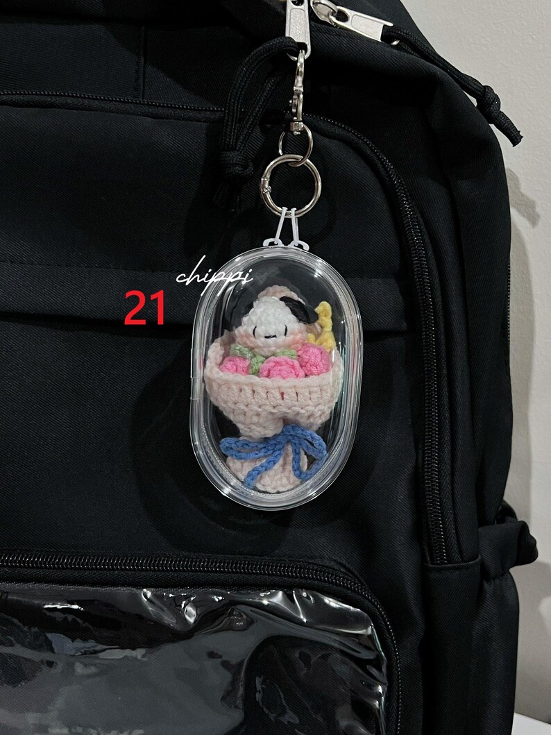 May include: A clear, oval keychain featuring a crocheted Snoopy character with a bouquet of pink flowers, tied with a blue ribbon. The keychain is attached to a black backpack. The number "21" and the word "chippi" are printed on the keychain.
