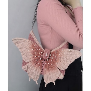 May include: A crochet butterfly-shaped handbag in shades of pink and cream, adorned with faux pearls. The bag features a chain strap and a button closure. The wings have a gradient effect, transitioning from a darker pink to a lighter cream color.