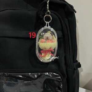 May include: A clear, oval keychain featuring a small crocheted dog and flowers. The keychain is attached to a black backpack. The number "19" is visible on the backpack. The word "chippi" is written on the keychain.