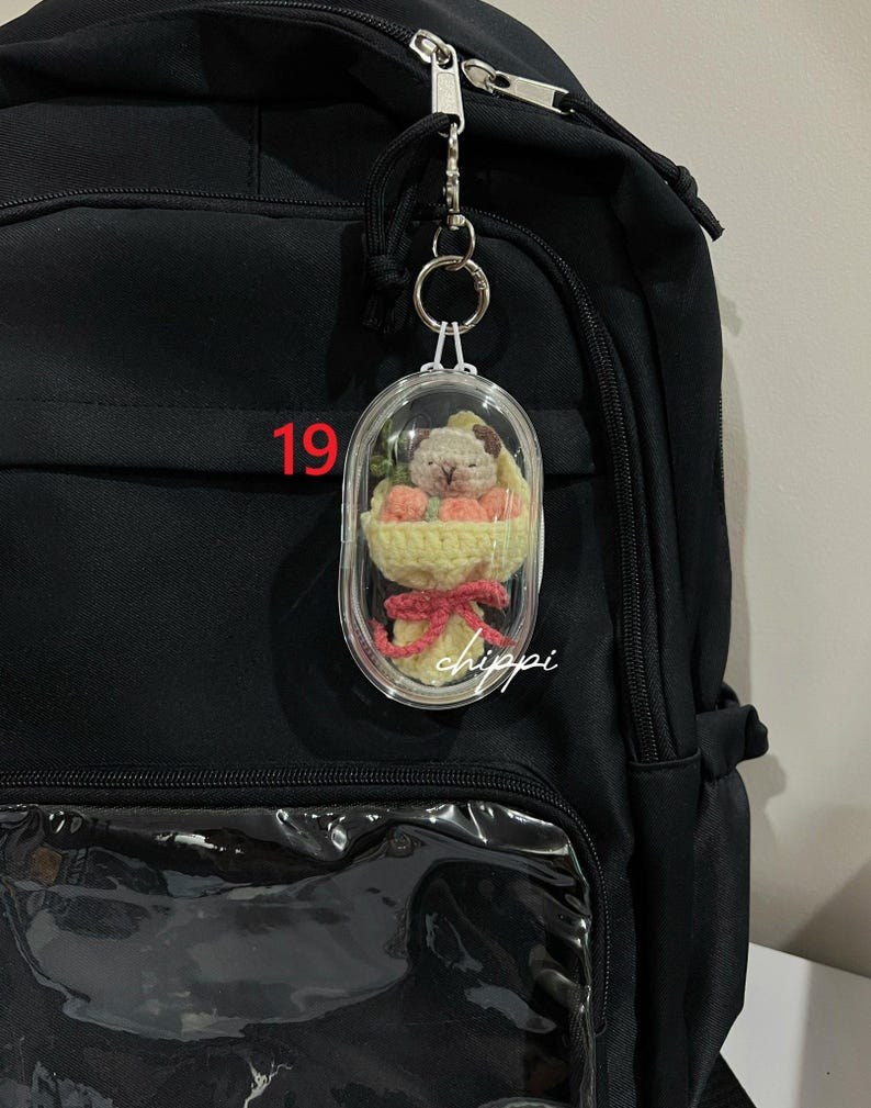 May include: A clear, oval keychain featuring a small crocheted dog and flowers. The keychain is attached to a black backpack. The number "19" is visible on the backpack. The word "chippi" is written on the keychain.