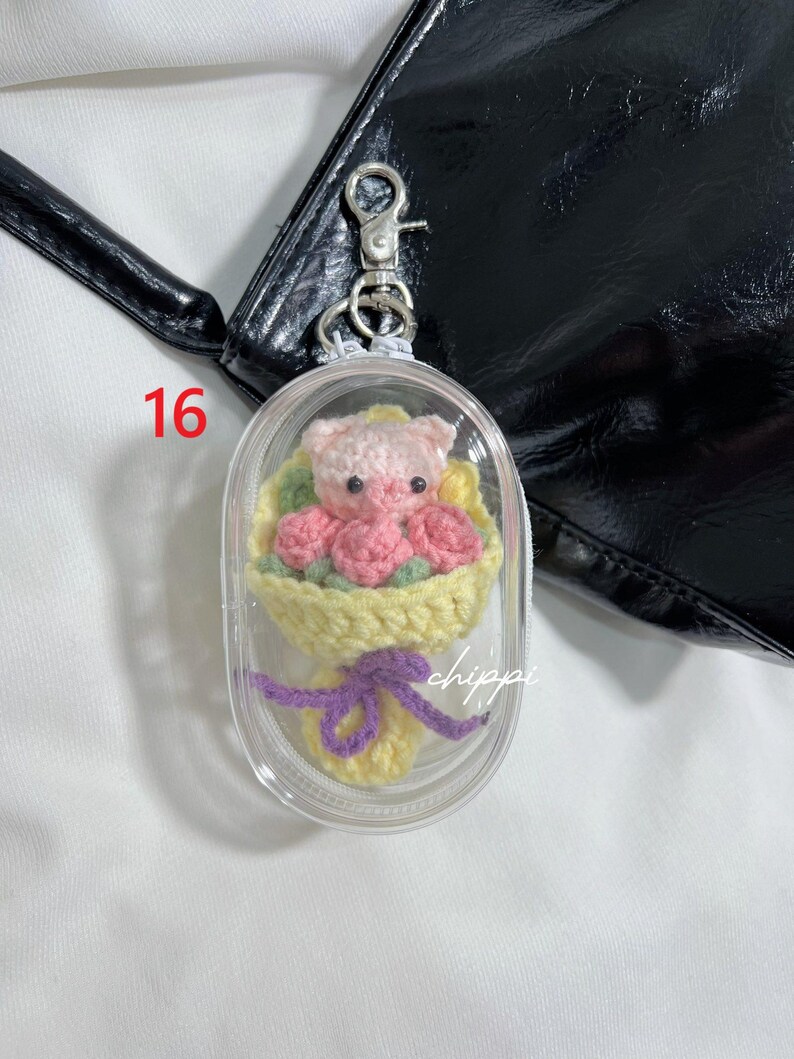 May include: A clear keychain featuring a crocheted bouquet and a pink pig. The bouquet has pink roses and yellow wrapping, tied with a purple bow. The number 16 is in red. The word "chippi" is visible.