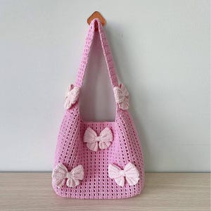 May include: A pink crocheted tote bag with four pink bows. The bag has a single strap and is hanging from a hook.