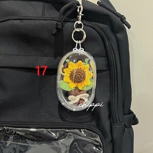 May include: A clear, oval keychain featuring a crocheted sunflower, attached to a black backpack. The sunflower has yellow petals and a brown centre. The number "17" is visible on the backpack, and the word "hippi" is written in cursive.