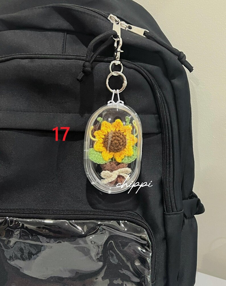 May include: A clear, oval keychain featuring a crocheted sunflower, attached to a black backpack. The sunflower has yellow petals and a brown centre. The number "17" is visible on the backpack, and the word "hippi" is written in cursive.