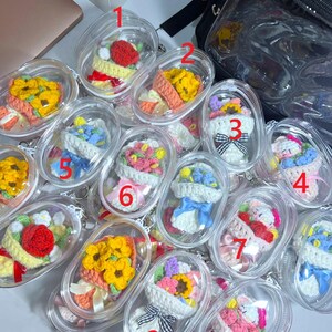 May include: A selection of crochet flower bouquets presented in clear plastic containers. The bouquets display a variety of colours, such as yellow, red, pink, and blue. Each bouquet is carefully crafted with yarn and ribbon details, demonstrating a handmade quality.