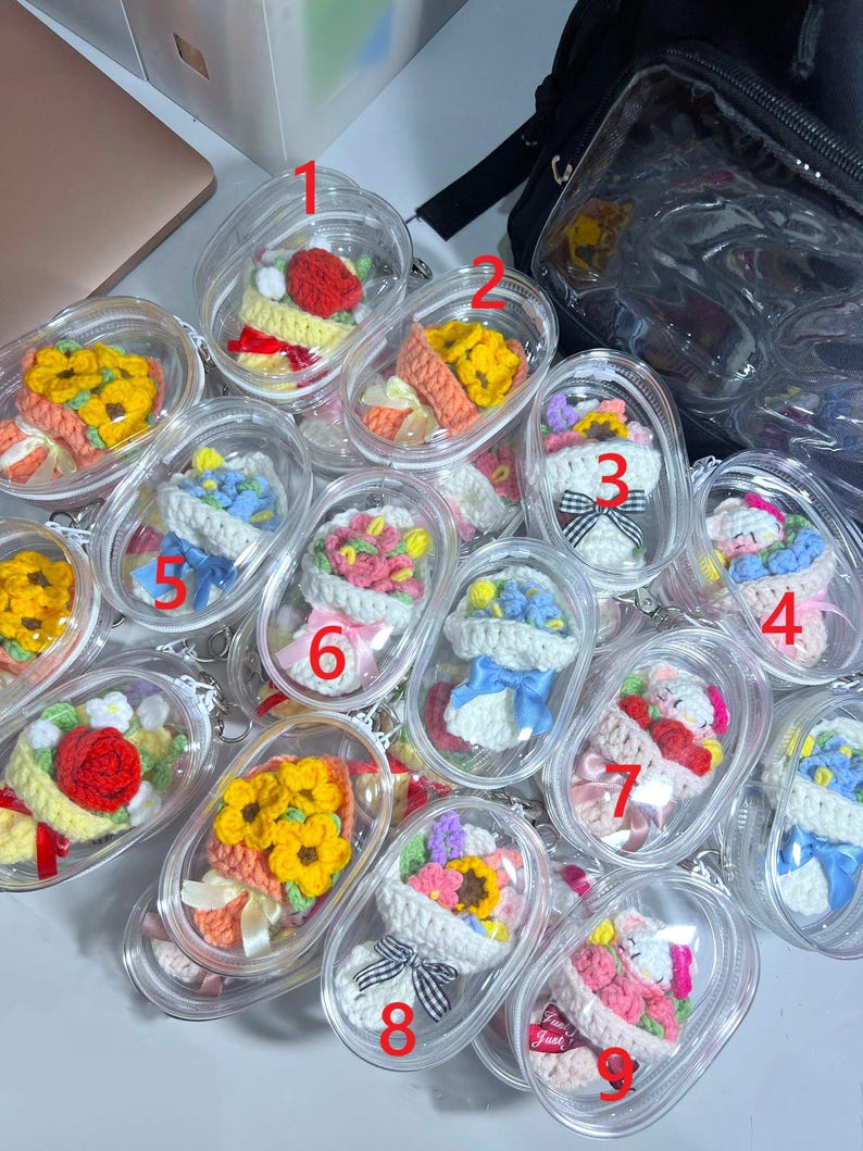 May include: A selection of crochet flower bouquets presented in clear plastic containers. The bouquets display a variety of colours, such as yellow, red, pink, and blue. Each bouquet is carefully crafted with yarn and ribbon details, demonstrating a handmade quality.