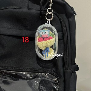 May include: A clear, oval keychain featuring a crocheted bouquet of red flowers and a blue-faced character. The bouquet is wrapped in yellow yarn and tied with a blue ribbon. The number "18" is visible in red. The word "chippi" is written in white.