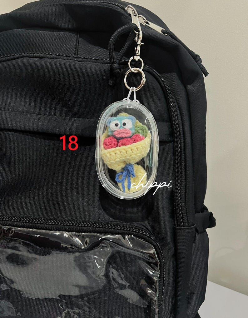 May include: A clear, oval keychain featuring a crocheted bouquet of red flowers and a blue-faced character. The bouquet is wrapped in yellow yarn and tied with a blue ribbon. The number "18" is visible in red. The word "chippi" is written in white.