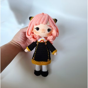 May include: A handmade crochet doll with pink hair, black and yellow cat ears, and a black dress with gold accents. The doll has a white collar with a red bow and black shoes. The doll is approximately 8 inches tall.
