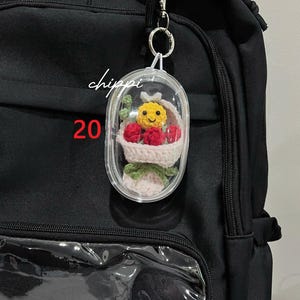May include: A black backpack with a clear pocket and a decorative keychain. The keychain features a small, clear capsule containing a crocheted basket with a yellow smiley face, red berries, and green leaves. The word "chippi" and the number "20" are visible.