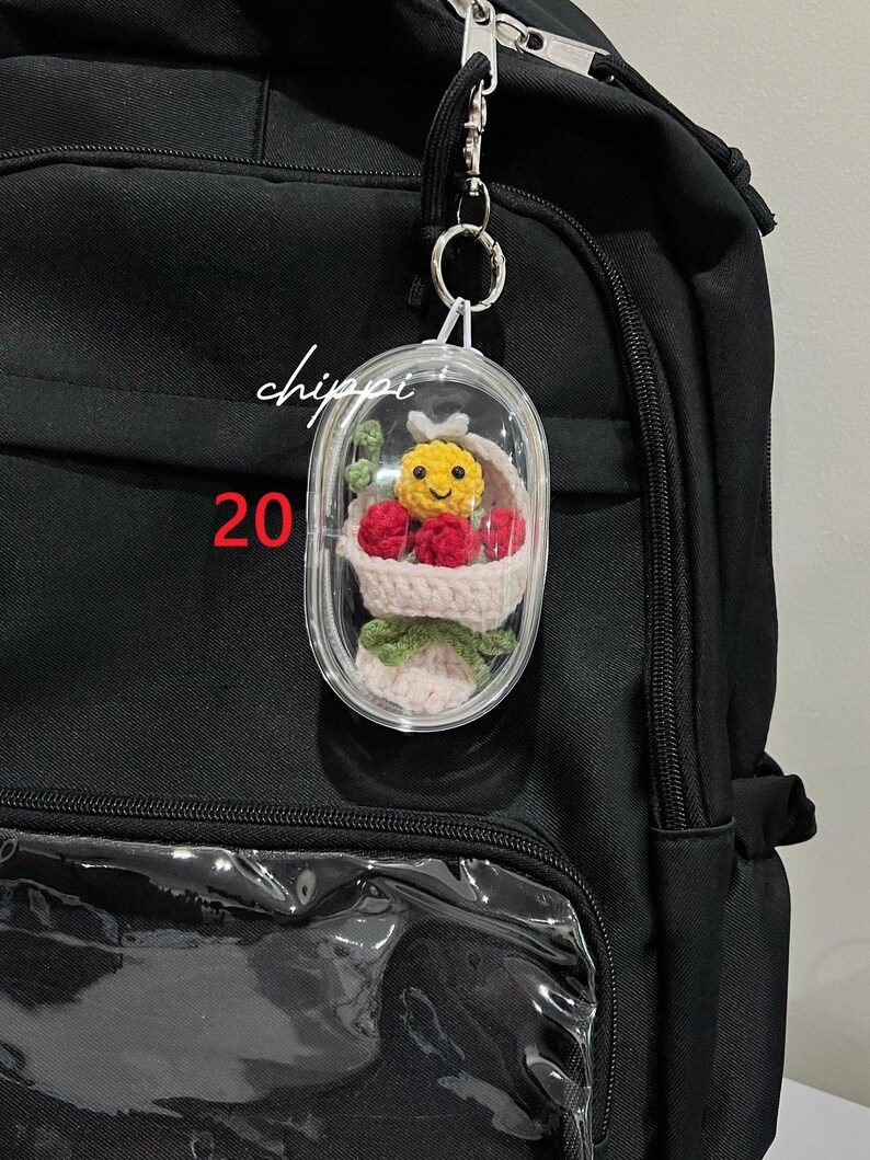 May include: A black backpack with a clear pocket and a decorative keychain. The keychain features a small, clear capsule containing a crocheted basket with a yellow smiley face, red berries, and green leaves. The word "chippi" and the number "20" are visible.
