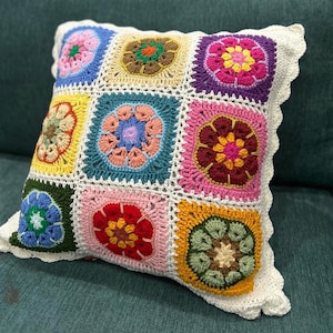 Könnte beinhalten: Ein quadratisches Häkelkissen mit Patchwork-Design. Das Kissen hat neun farbenfrohe Quadrate, jedes mit einem Blumenmotiv in Rosa-, Blau-, Gelb-, Grün- und Lilatönen. Die Kanten sind mit einem weißen, gewellten Rand versehen.