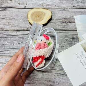 May include: A clear phone case containing a crocheted bouquet. The bouquet includes a white character with a red bow, pink flowers, and a red bow. The case is held open, showcasing the handmade craft within. The case is made of plastic.