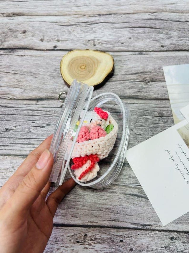 May include: A clear phone case containing a crocheted bouquet. The bouquet includes a white character with a red bow, pink flowers, and a red bow. The case is held open, showcasing the handmade craft within. The case is made of plastic.