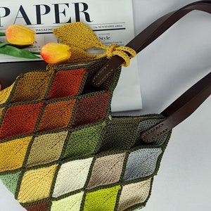 Puede incluir: Un bolso de mano de crochet con un patrón de rombos en tonos de verde, marrón, amarillo y gris. El bolso tiene asas de cuero marrón.