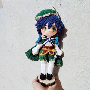 May include: A crocheted doll with blue hair, green clothing, and a green hat. The doll is standing on a round base with a sparkly design.