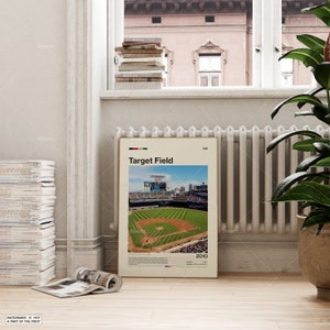 Op de afbeelding: Een ingelijste poster met Target Field honkbalstadion, met de tekst "Target Field" en "2010". De afbeelding toont het honkbalveld met groen gras, blauwe lucht en stadionzitplaatsen. De poster staat in een kamer met een raam en een radiator.