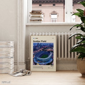 May include: A framed poster featuring Soldier Field, a stadium in Chicago, Illinois. The poster has a cream-colored background with the stadium's name and a colorful graphic at the top. The image shows the stadium at dusk, with the city skyline in the background.
