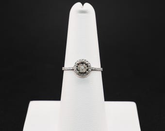 May include: A silver ring with a central cluster of small stones surrounded by a halo of smaller stones. The ring is set on a white display stand against a black background. The band is also adorned with small stones.