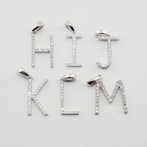 May include: Six silver-toned metal letter pendants shaped as H, I, J, K, L, and M. Each letter is encrusted with small, clear, sparkling stones and features a polished metal bail at the top for attachment to a chain or necklace cord.