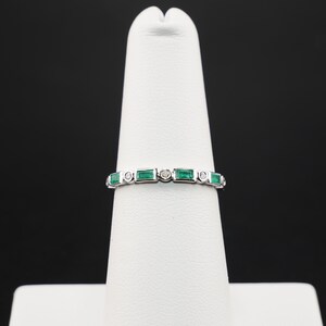 May include: A silver ring with alternating emerald-cut green gemstones and small round clear gemstones. The ring is displayed on a white stand against a black background. The gemstones are set in a continuous band.