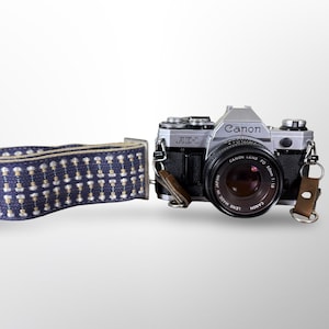 May include: A silver Canon AE-1 camera with a black lens and a blue and white patterned camera strap. The camera strap is made of woven fabric. The camera lens reads "CANON LENS FD 50mm 1:1.8".