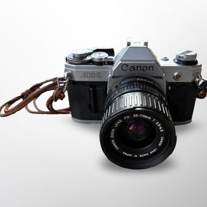 Puede incluir: Una cámara Canon AE-1 plateada y negra con un objetivo zoom. El objetivo es negro con detalles plateados y tiene el texto "35-70mm 1:3.5-4.5" impreso. Una correa de cuero marrón está sujeta a la cámara.