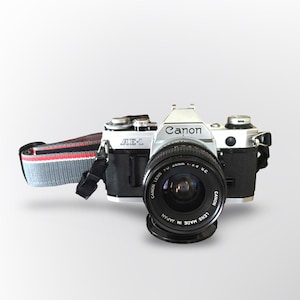 Puede incluir: Una cámara vintage Canon AE-1 con cuerpo negro y parte superior plateada. La lente de la cámara es negra con las palabras "Canon Lens Made in Japan" impresas. La correa de la cámara es gris, roja y negra.