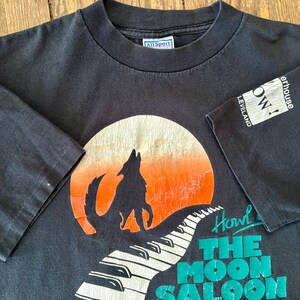 May include: Black t-shirt with a graphic of a wolf howling at the moon, a piano keyboard, and the text "Howl at The Moon Saloon". The shirt has a white tag with the text "All Sport" and a white patch with the text "The Warehouse Cleveland, OH!"