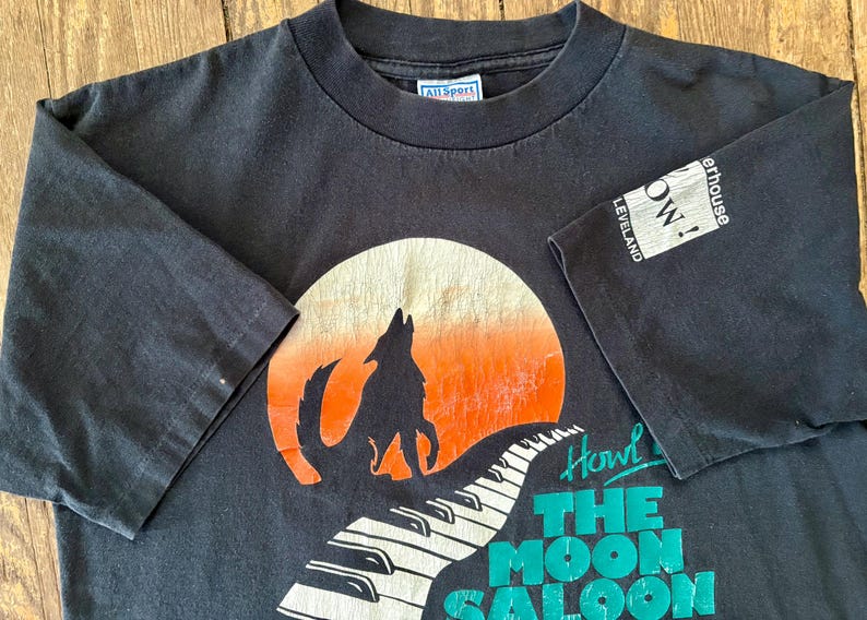 May include: Black t-shirt with a graphic of a wolf howling at the moon, a piano keyboard, and the text "Howl at The Moon Saloon". The shirt has a white tag with the text "All Sport" and a white patch with the text "The Warehouse Cleveland, OH!"