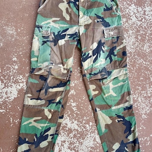 May include: Cargo trousers featuring a woodland camouflage pattern in shades of green, brown, and black. These trousers include two large side cargo pockets with button flaps, a button fly closure, and adjustable side waist tabs for a secure fit.