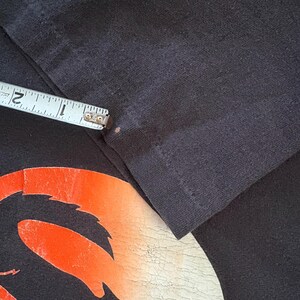 May include: Close-up of a black t-shirt with a red and orange graphic design. The graphic design is a stylized image of a creature with a long tail and sharp teeth.