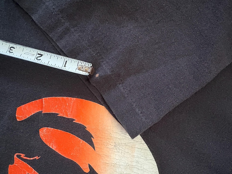 May include: Close-up of a black t-shirt with a red and orange graphic design. The graphic design is a stylized image of a creature with a long tail and sharp teeth.