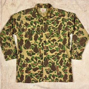 May include: A long-sleeve button-down shirt featuring a camouflage pattern in shades of tan, brown, and green. The shirt includes a pointed collar, two chest pockets with buttoned flaps, and a front button closure. A label reading Kamat is visible.