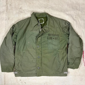 May include: An olive green jacket featuring a pointed collar, a button-down front closure, and a single chest pocket with a button flap. The garment includes two lower side pockets and small adjustable side tabs at the hemline.