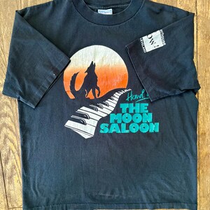 May include: Black t-shirt with a graphic of a wolf howling at a moon with a piano keyboard in the foreground. The text "Howl at The Moon Saloon" is printed on the shirt.