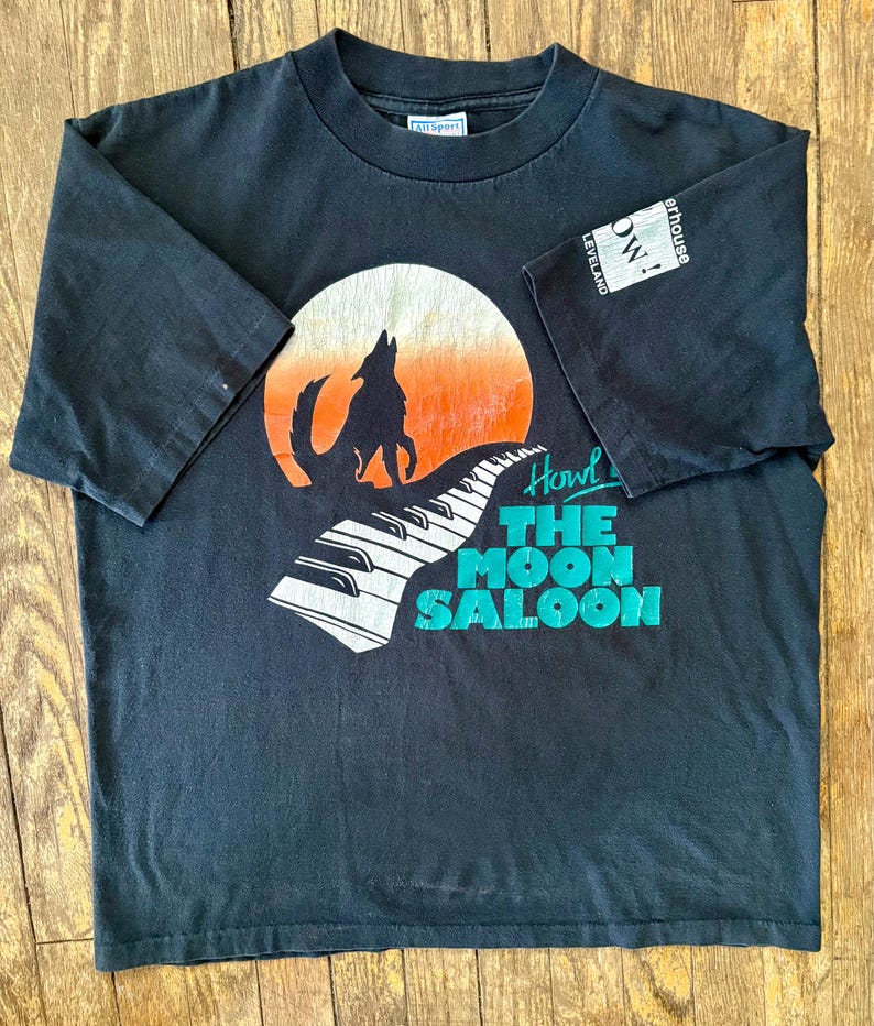 May include: Black t-shirt with a graphic of a wolf howling at a moon with a piano keyboard in the foreground. The text "Howl at The Moon Saloon" is printed on the shirt.