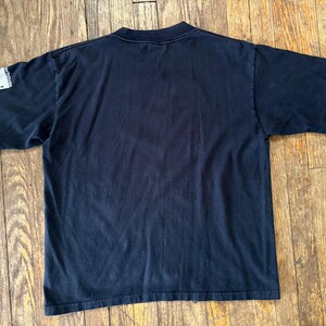 May include: Black short-sleeved t-shirt with a white patch on the left sleeve that says "NOW!" in all caps.