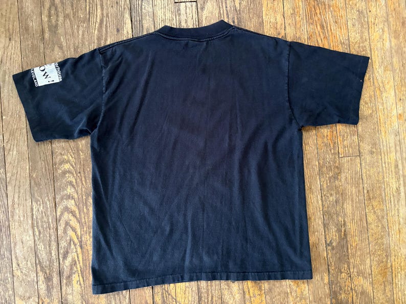 May include: Black short-sleeved t-shirt with a white patch on the left sleeve that says "NOW!" in all caps.