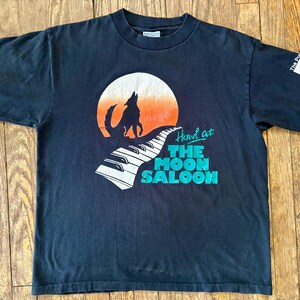 May include: Black t-shirt with a graphic of a wolf howling at the moon with a piano keyboard in the foreground. The text "Howl at The Moon Saloon" is printed below the graphic.