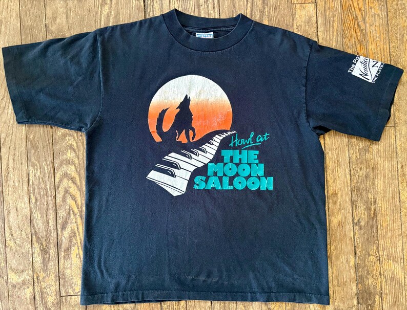 May include: Black t-shirt with a graphic of a wolf howling at the moon with a piano keyboard in the foreground. The text "Howl at The Moon Saloon" is printed below the graphic.