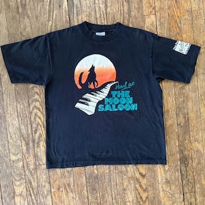 May include: Black t-shirt with a graphic of a wolf howling at the moon, a piano keyboard, and the text "Howl at The Moon Saloon".