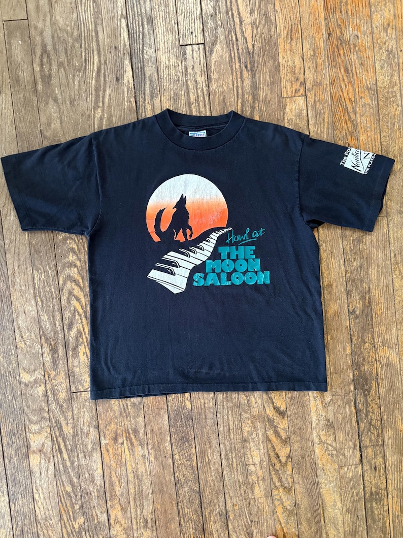 May include: Black t-shirt with a graphic of a wolf howling at the moon, a piano keyboard, and the text "Howl at The Moon Saloon".