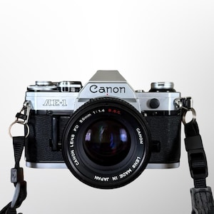 May include: A Canon AE-1 camera with a black lens and strap. The camera body is silver and black, with the Canon logo displayed. The lens reads "CANON LENS FD 50mm 1:1.4 S.S.C." and "LENS MADE IN JAPAN."